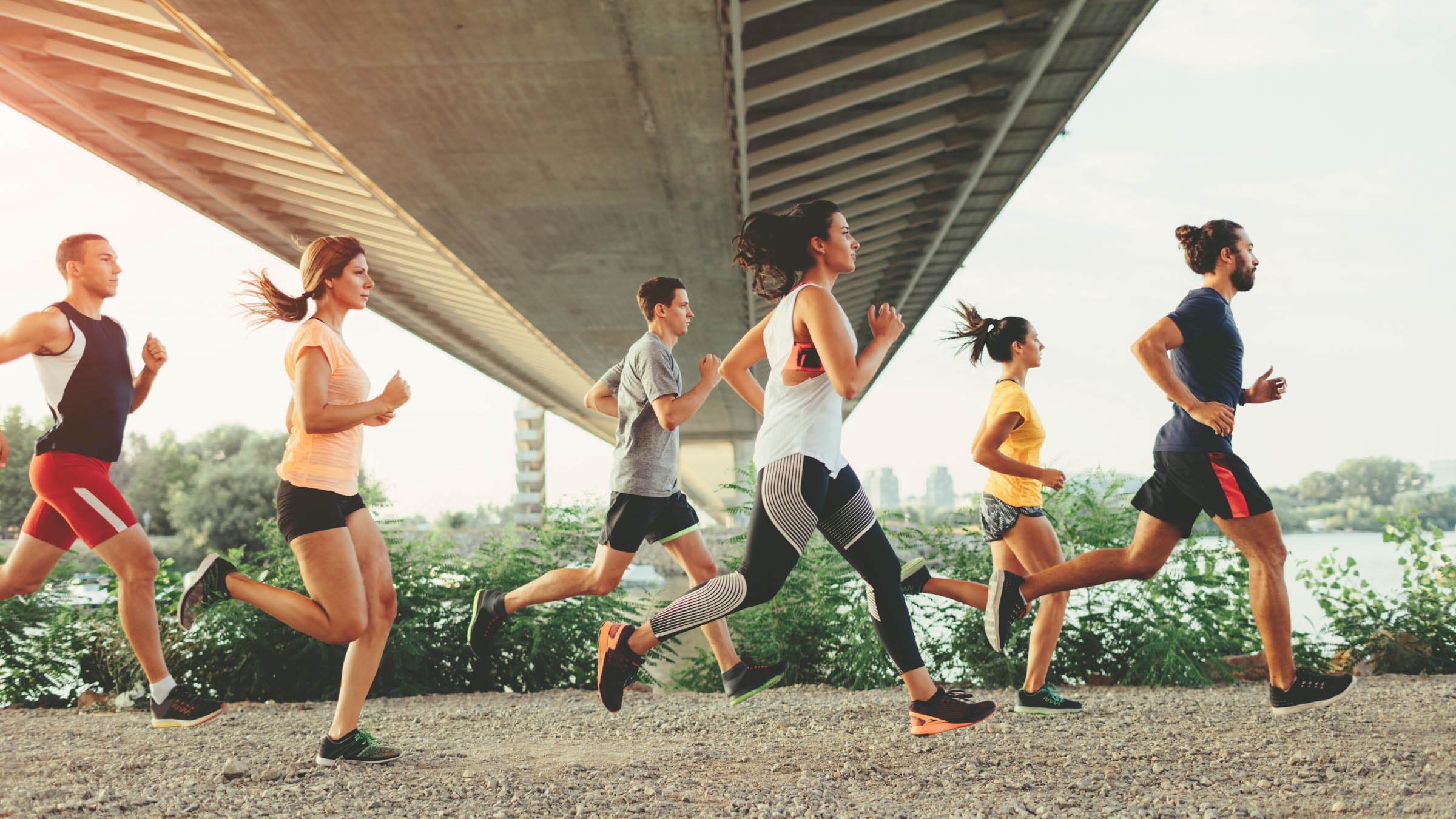 Side view of group of runners running under the bridge near river at sunset.